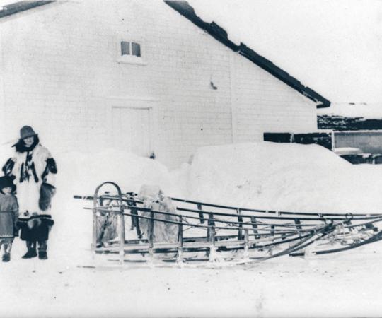 A woman and child stand in front of the Bone House on Herschel Island, where two Inuit men were hanged for murder in 1924. PWNHC: N-1991-041-0065